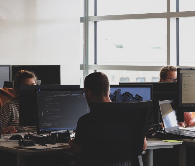 People working at desks with multiple computer monitors in an office with large windows.