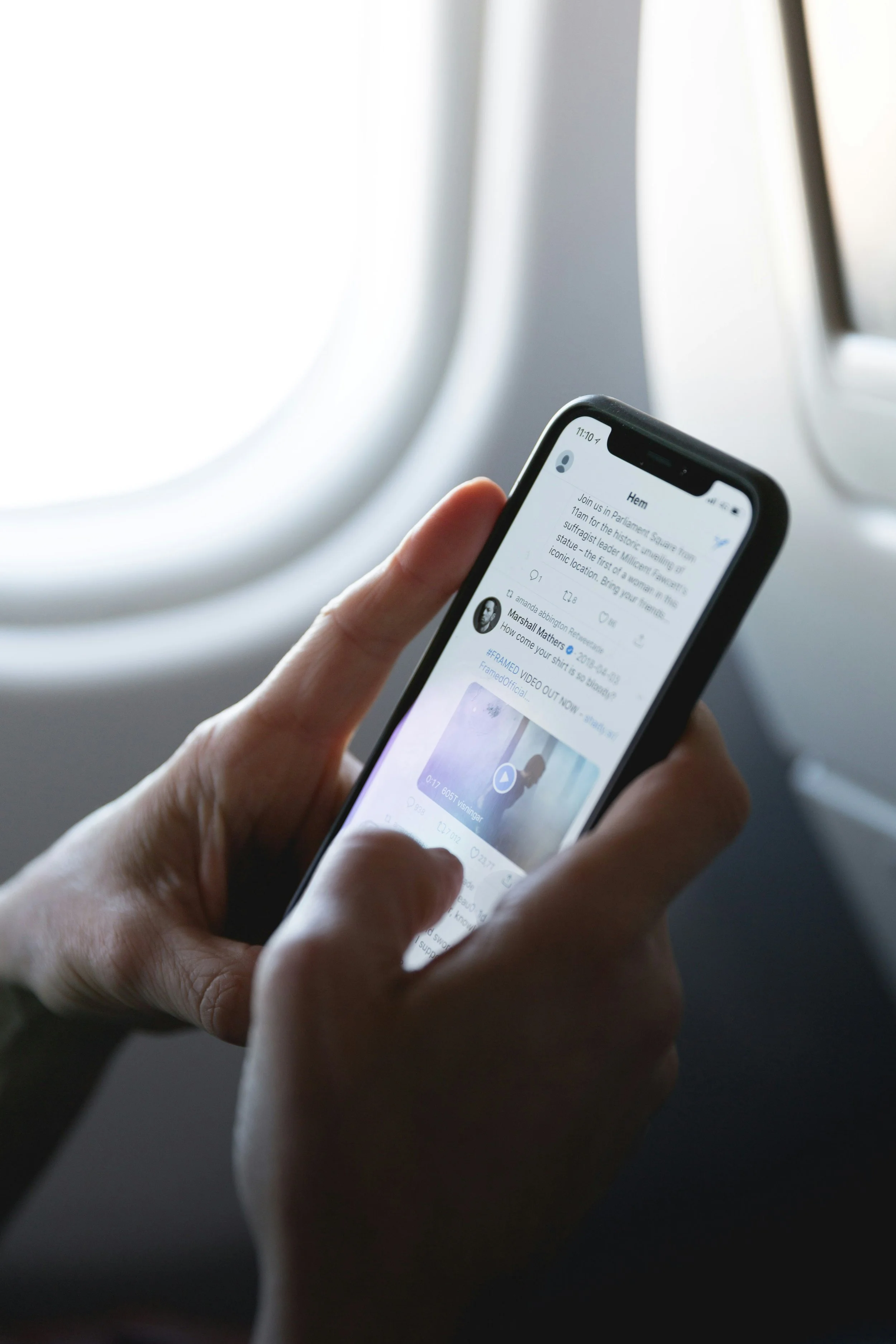 Person holding a smartphone displaying a social media feed, seated near an airplane window.