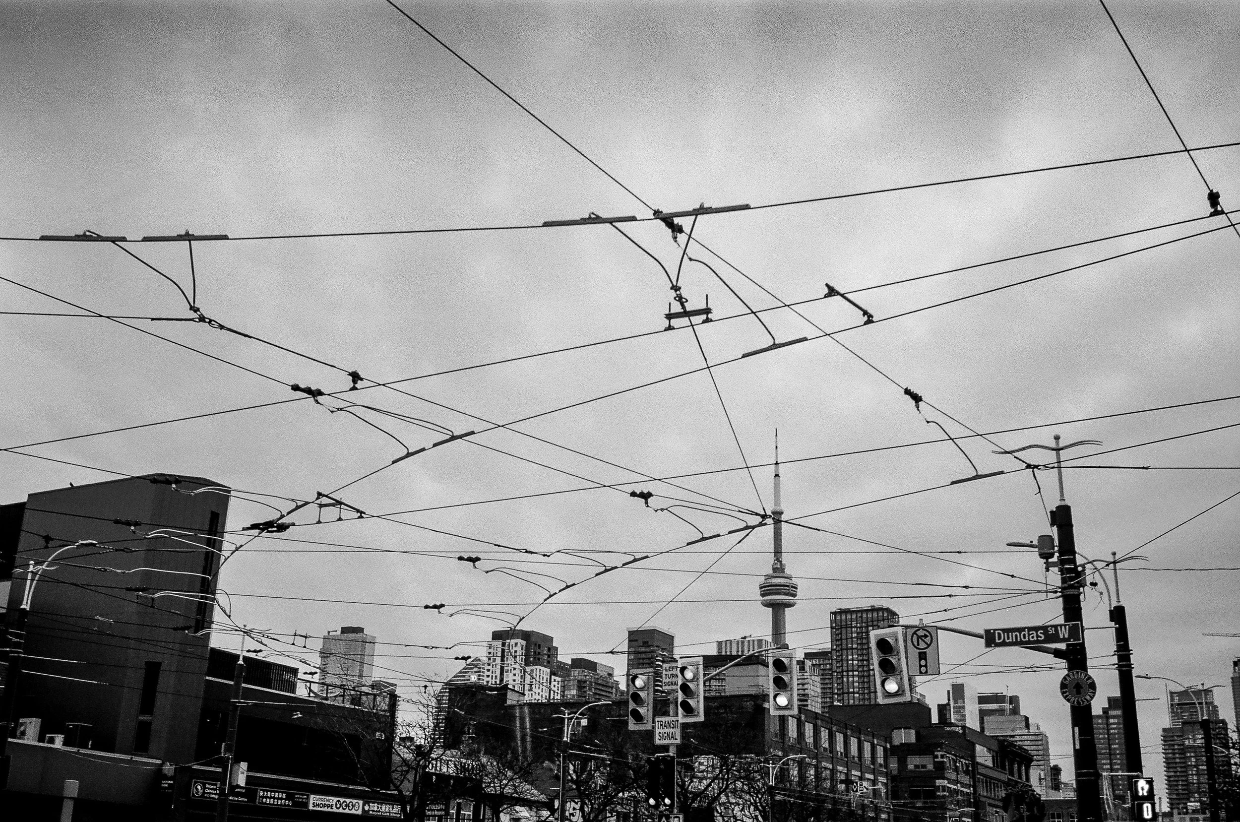 Black and white cityscape with overhead tram wires, traffic signals, and the CN Tower in the background in Toronto, Canada.