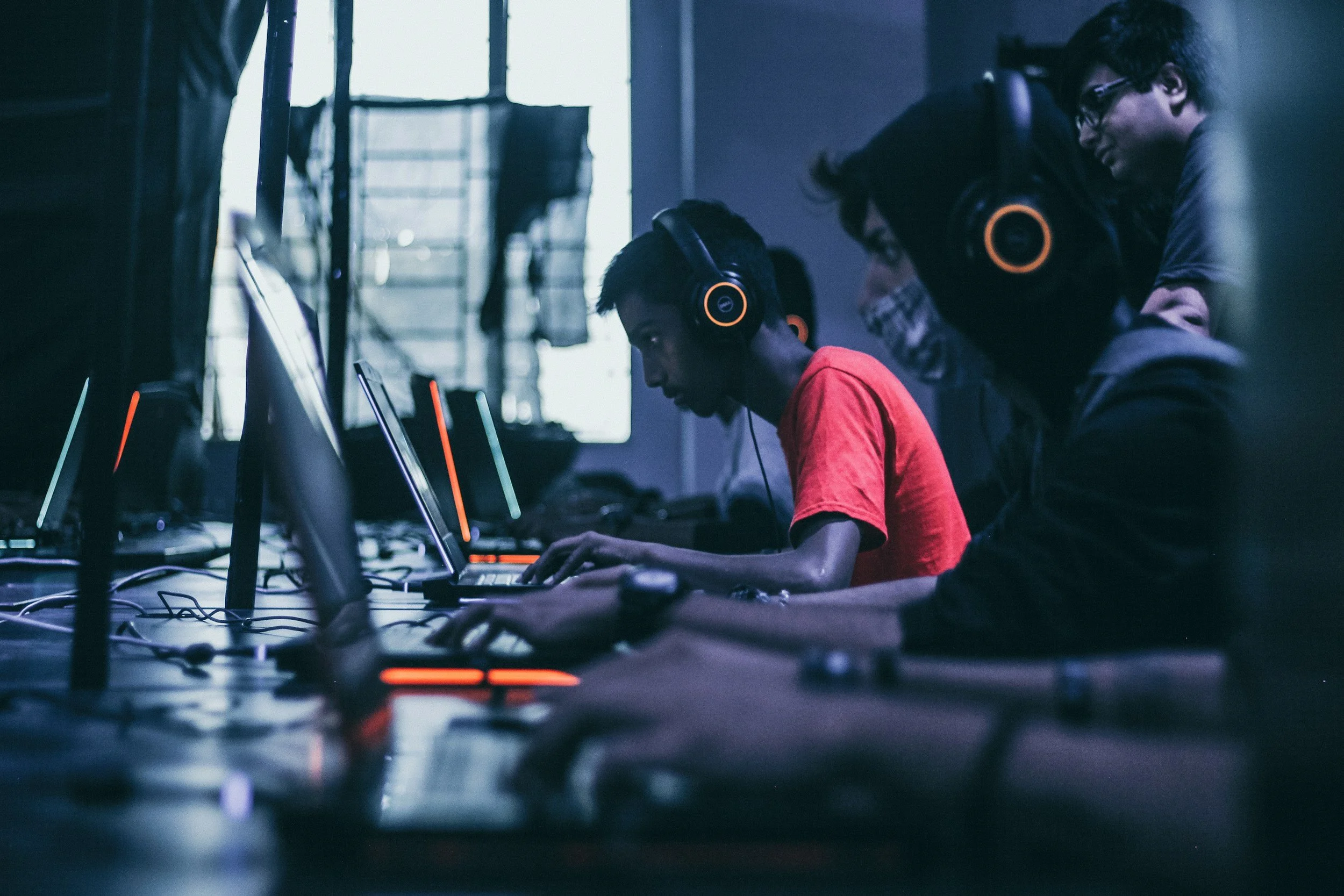 Group of young people playing computer games in a gaming cafe, wearing headphones, focused on their screens.